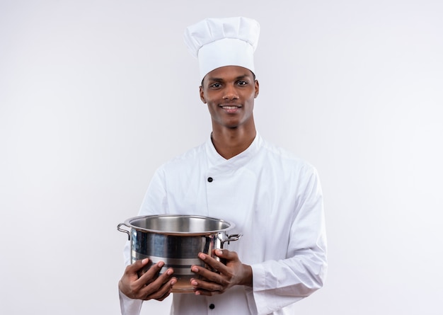 Sonriente joven cocinero afroamericano en uniforme de chef sostiene una cacerola y mira a la cámara sobre fondo blanco aislado con espacio de copia