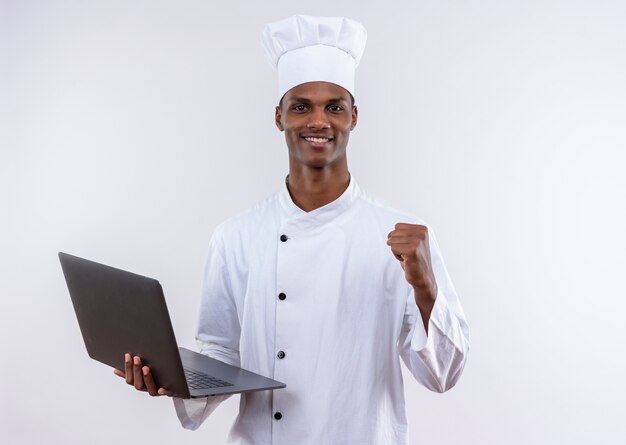 Sonriente joven cocinero afroamericano en uniforme de chef sostiene portátil y mantiene el puño en alto sobre fondo blanco aislado con espacio de copia