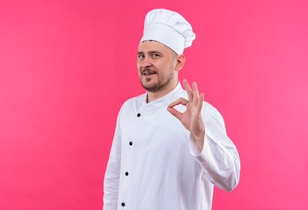 Sonriente joven cocinero guapo en uniforme de chef haciendo bien signo aislado en espacio rosa