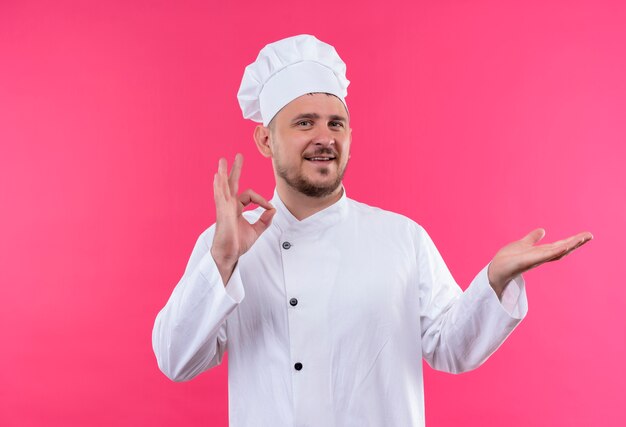 Sonriente joven cocinero guapo en uniforme de chef haciendo el signo de ok y mostrando la mano vacía aislada en el espacio rosa