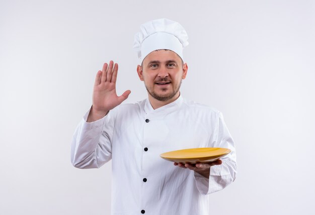 Sonriente joven cocinero guapo en uniforme de chef sosteniendo la placa y gesticulando hola aislado en el espacio en blanco