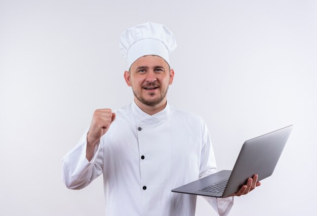 Sonriente joven cocinero guapo en uniforme de chef sosteniendo el portátil y levantando el puño en un espacio en blanco aislado