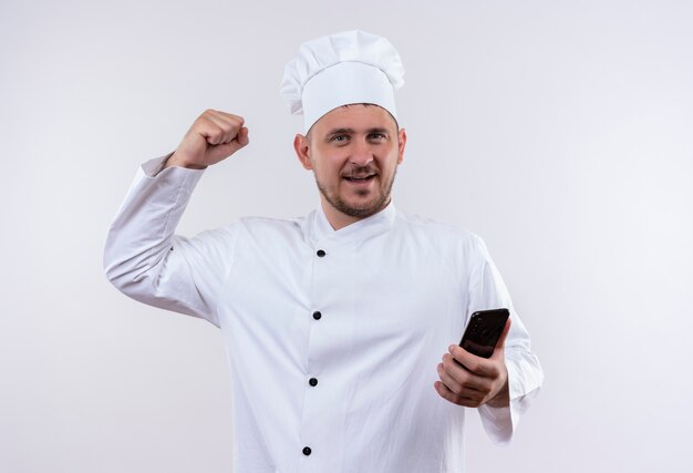 Sonriente joven cocinero guapo en uniforme de chef sosteniendo el teléfono móvil y haciendo un gesto fuerte aislado en el espacio en blanco