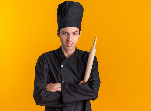 Sonriente joven cocinero rubio en uniforme de chef y gorra de pie con postura cerrada sosteniendo rodillo mirando a cámara aislada en pared naranja con espacio de copia