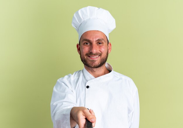 Sonriente joven cocinero de sexo masculino caucásico en uniforme de chef y gorra mirando a la cámara estirando la sartén hacia la cámara aislada en la pared verde oliva