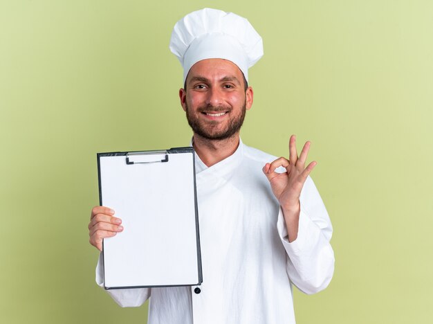 Sonriente joven cocinero de sexo masculino caucásico en uniforme de chef y gorra mirando a cámara mostrando portapapeles haciendo bien signo aislado en la pared verde oliva