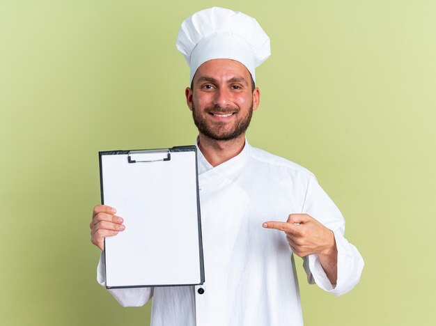 Sonriente joven cocinero de sexo masculino caucásico en uniforme de chef y gorra mostrando y apuntando al portapapeles