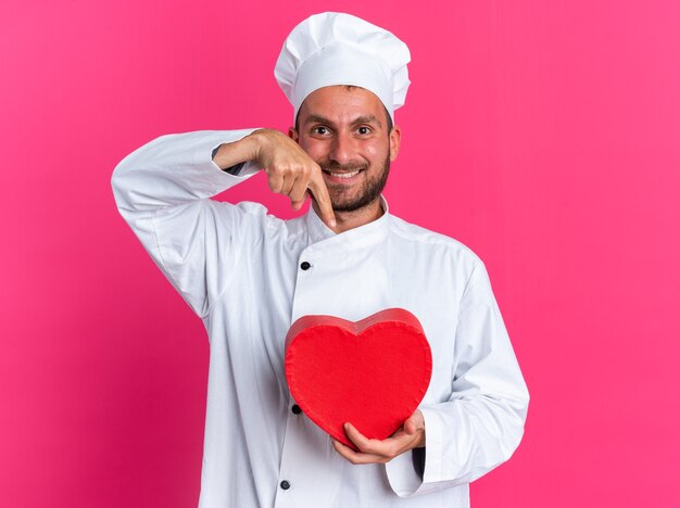 Sonriente joven cocinero de sexo masculino caucásico en uniforme de chef y gorra sosteniendo y apuntando a la forma del corazón mirando a cámara aislada en la pared rosa