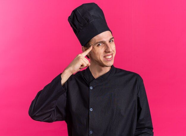 Sonriente joven cocinero de sexo masculino rubio en uniforme de chef y gorra haciendo gesto de pensar