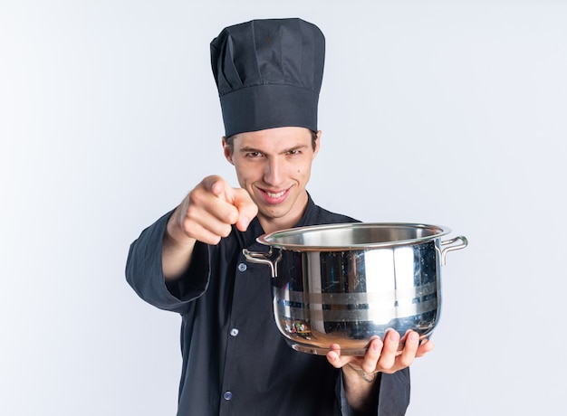 Sonriente joven cocinero de sexo masculino rubio en uniforme de chef y gorra mirando y apuntando a la cámara estirando la olla hacia la cámara aislada en la pared blanca