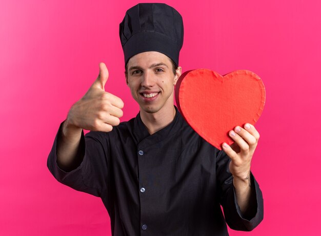 Sonriente joven cocinero de sexo masculino rubio en uniforme de chef y gorra mirando a cámara mostrando forma de corazón y pulgar hacia arriba aislado en la pared rosa