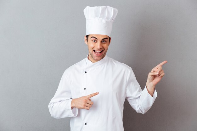 Sonriente joven cocinero en uniforme apuntando.