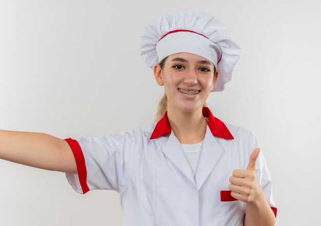 Sonriente joven cocinero en uniforme de chef con aparatos dentales y brazo abierto mostrando el pulgar hacia arriba aislado en el espacio en blanco