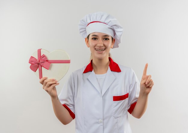 Sonriente a joven cocinero en uniforme de chef con aparatos dentales sosteniendo una caja de regalo en forma de corazón y levantando el dedo aislado en el espacio en blanco