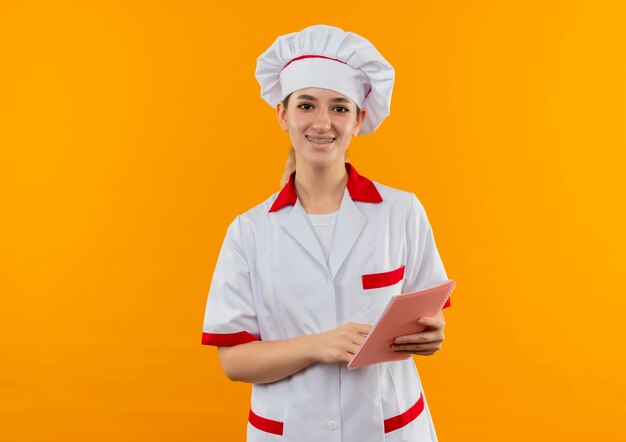 Sonriente joven cocinero en uniforme de chef con aparatos dentales sosteniendo y poniendo el dedo en el bloc de notas aislado en el espacio naranja