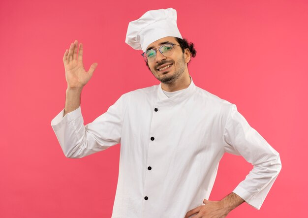 Sonriente joven cocinero con uniforme de chef y gafas levantando la mano y poniendo otra mano en la cadera en rosa