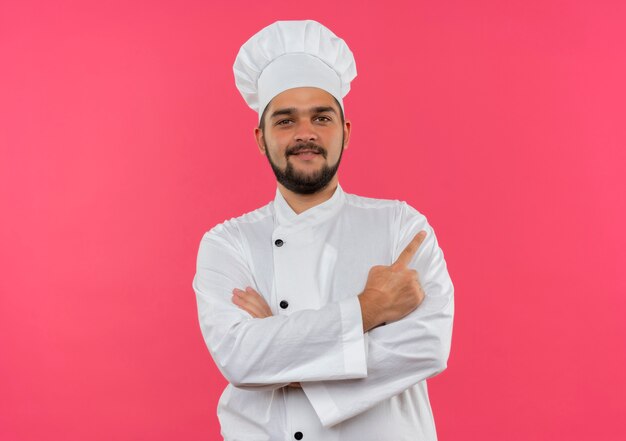 Sonriente joven cocinero en uniforme de chef de pie con una postura cerrada y apuntando al lado aislado en el espacio rosa
