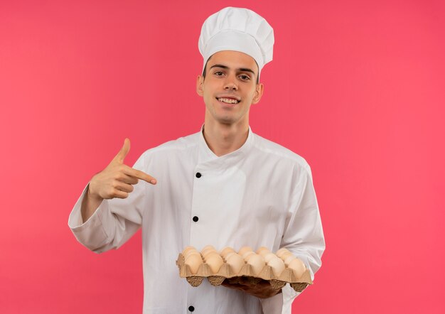Sonriente joven cocinero vistiendo uniforme de chef señala con el dedo al lote de huevos en su mano con espacio de copia