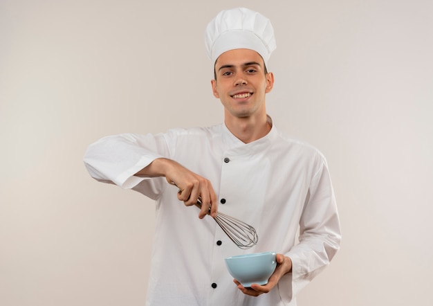 Sonriente joven cocinero vistiendo uniforme de chef sosteniendo batidor con tazón