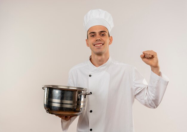 Sonriente joven cocinero vistiendo uniforme de chef sosteniendo una cacerola mostrando sí gesto