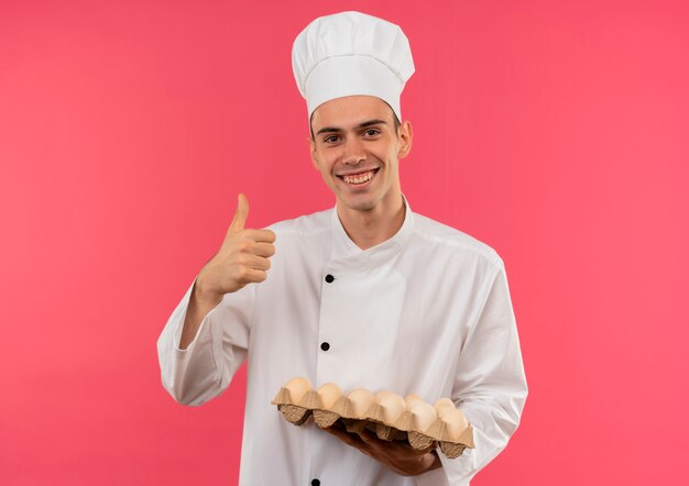 Sonriente joven cocinero vistiendo uniforme de chef sosteniendo un lote de huevos con el pulgar hacia arriba