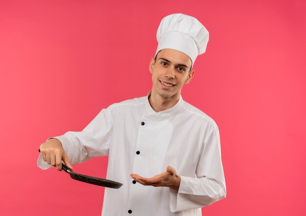 Sonriente joven cocinero vistiendo uniforme de chef sosteniendo y puntos sartén con espacio de copia