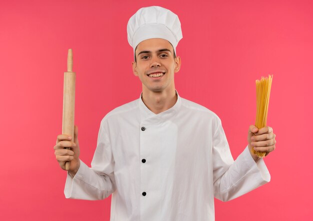 Sonriente joven cocinero vistiendo uniforme de chef sosteniendo un rodillo y espaguetis