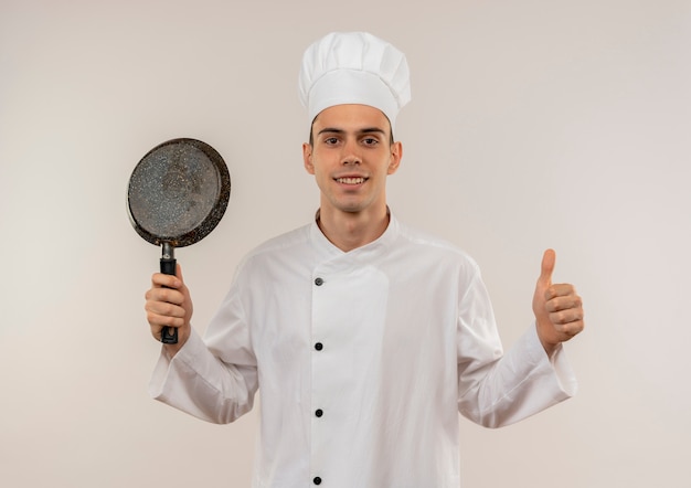 Sonriente joven cocinero vistiendo uniforme de chef sosteniendo la sartén con el pulgar hacia arriba