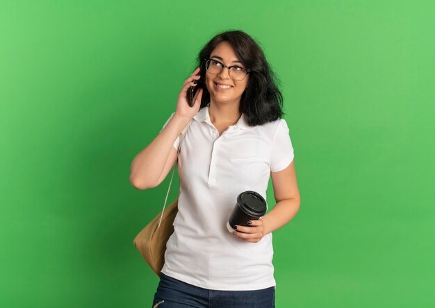 Sonriente joven colegiala bastante caucásica con gafas y bolsa trasera habla por teléfono sosteniendo la taza de café en verde con espacio de copia