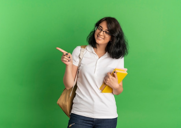 Sonriente joven colegiala bastante caucásica con gafas y mochila mira y apunta al lado sosteniendo libros en verde con espacio de copia