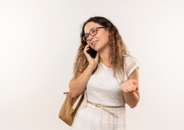 Sonriente joven colegiala bonita con gafas y bolsa trasera mostrando la mano vacía y hablando por teléfono mirando al lado aislado en la pared