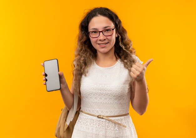 Sonriente joven colegiala bonita con gafas y bolsa trasera que muestra el teléfono móvil y el pulgar hacia arriba aislado en la pared amarilla