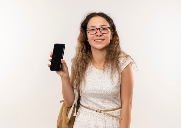 Sonriente joven colegiala bonita con gafas y bolso trasero mostrando teléfono móvil aislado en la pared blanca