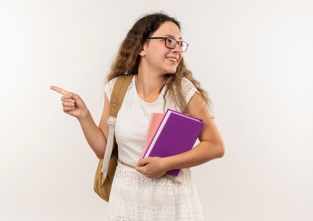 Sonriente joven colegiala bonita con gafas y mochila sosteniendo libro y bloc de notas apuntando y mirando al lado aislado en la pared blanca