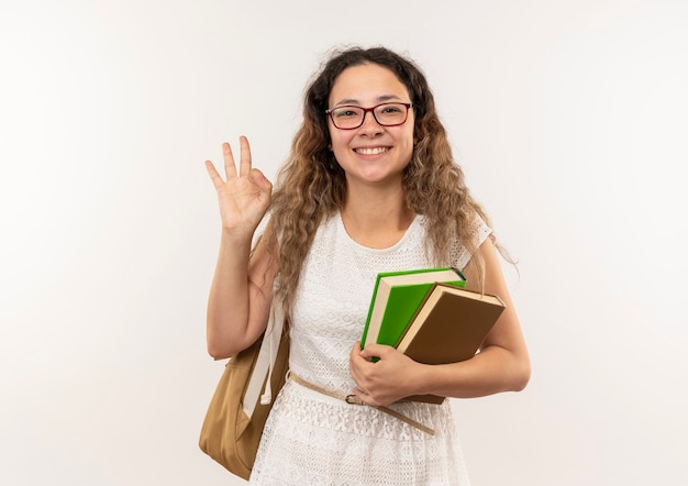 Foto gratuita sonriente joven colegiala bonita con gafas y mochila sosteniendo libros y haciendo bien firmar aislado en la pared blanca