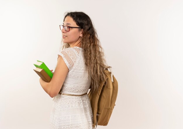 Sonriente joven colegiala bonita con gafas y mochila sosteniendo libros mirando al lado de pie detrás de la vista aislada en la pared blanca