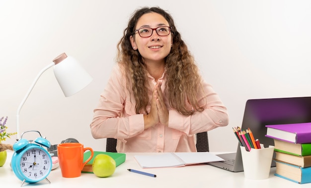 Sonriente joven colegiala bonita con gafas sentado en el escritorio con herramientas escolares haciendo sus deberes poniendo las manos en gesto de oración mirando hacia arriba aislado en la pared blanca