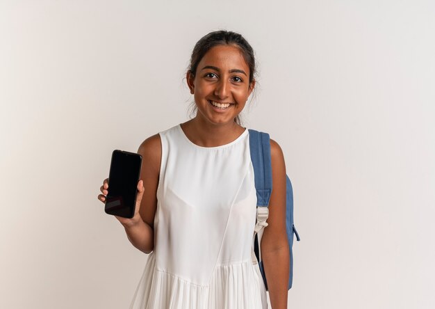 Sonriente joven colegiala vistiendo bolsa trasera sosteniendo el teléfono aislado en la pared blanca