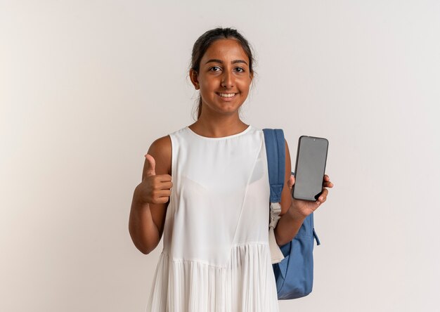 Sonriente joven colegiala vistiendo bolsa trasera sosteniendo el teléfono con el pulgar hacia arriba aislado en blanco