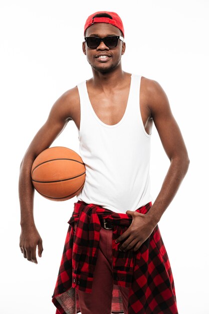 Sonriente joven confiado de pie y sosteniendo la pelota de baloncesto