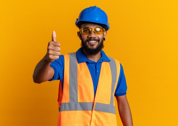 Sonriente joven constructor afroamericano en gafas de seguridad con uniforme con casco de seguridad pulgar hacia arriba aislado sobre fondo naranja con espacio de copia