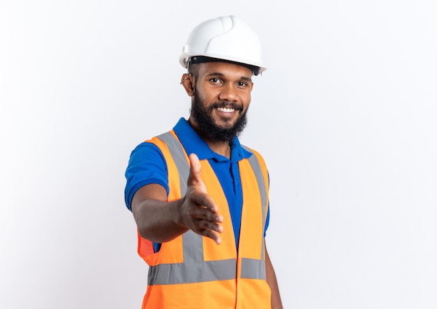 Sonriente joven constructor afroamericano en uniforme con casco de seguridad extendiendo su mano aislada sobre fondo blanco con espacio de copia