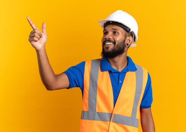 Sonriente joven constructor afroamericano en uniforme con casco de seguridad mirando y apuntando al lado aislado sobre fondo naranja con espacio de copia