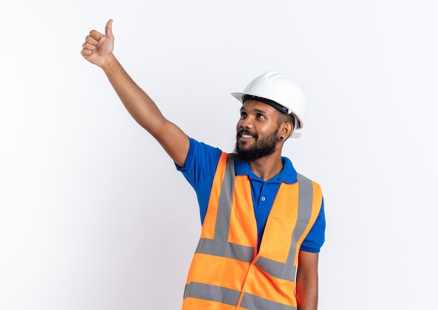 Sonriente joven constructor afroamericano en uniforme con casco de seguridad pulgar hacia arriba mirando al lado aislado sobre fondo blanco con espacio de copia