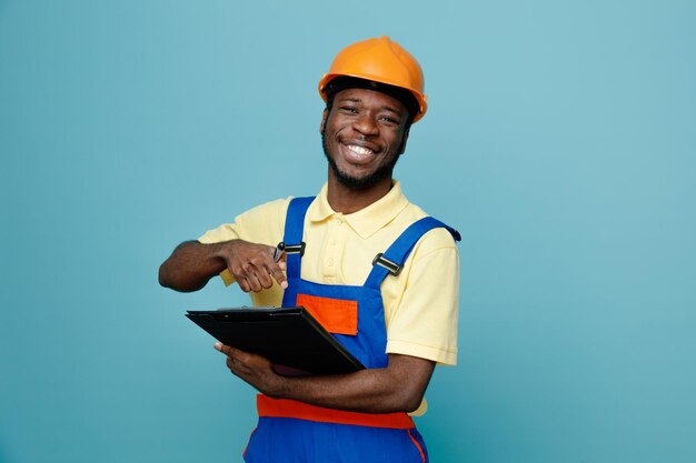 Sonriente joven constructor afroamericano en uniforme sosteniendo y apuntando al portapapeles aislado sobre fondo azul.