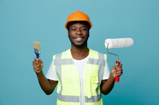 Sonriente joven constructor afroamericano en uniforme sosteniendo cepillo de rodillos con pincel aislado sobre fondo azul.