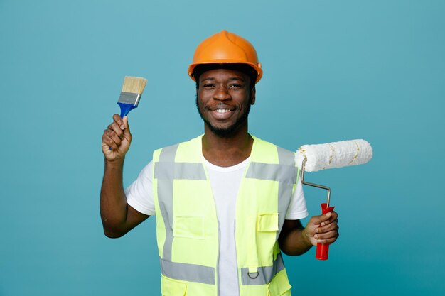 Sonriente joven constructor afroamericano en uniforme sosteniendo cepillo de rodillos con pincel aislado sobre fondo azul.