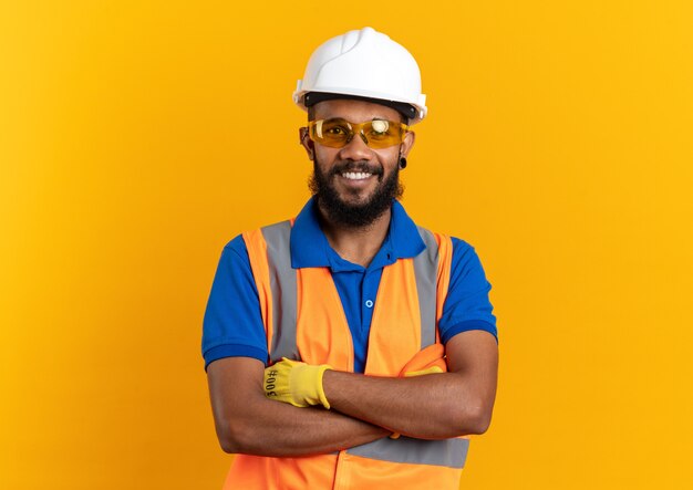 Sonriente joven constructor hombre en gafas de seguridad con uniforme con casco de seguridad de pie con los brazos cruzados aislado en la pared naranja con espacio de copia