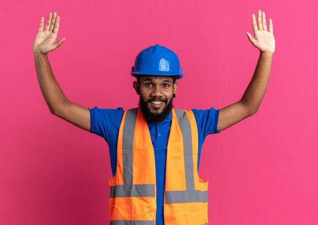 Sonriente joven constructor hombre en uniforme con casco de seguridad de pie con las manos levantadas aislado en la pared rosa con espacio de copia