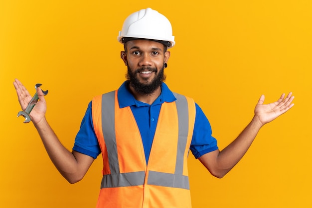 Sonriente joven constructor hombre en uniforme con casco de seguridad sosteniendo la llave del taller y manteniendo la mano abierta aislada en la pared naranja con espacio de copia